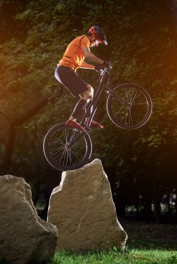 Extreme show of talented trail biker performing on big rocks standing on back wheel, in the evening in park, side view, concept of active lifestyle clipart