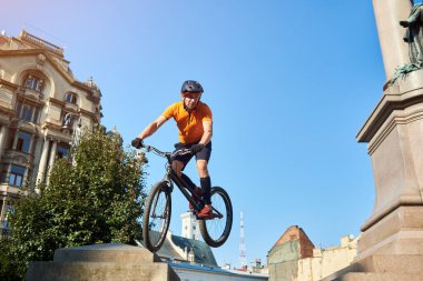 View from below athlete holding a bicycle in hands and looking away. Sportsman masterfully showing trick on bicycle on architecture background. Concept of active hobby.