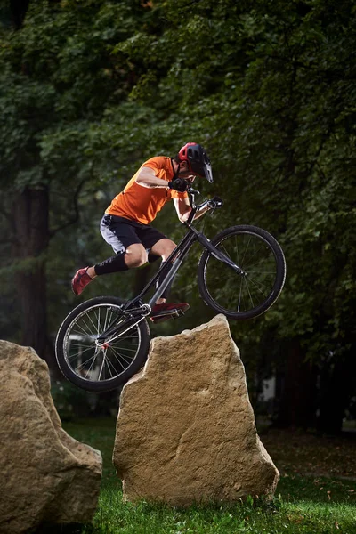 Front view of man holding a bicycle handlebar and doing a stunt over stones. Athlete trying to land on mountain bike on grass. Concept of land.