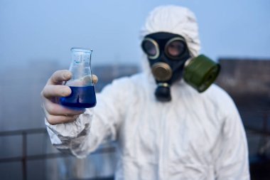 Elinde mavi sıvıyla cam bir şişe tutan, gaz maskesi takan ve koruyucu kıyafet giyen, kirlilik analizi yapan, yüksek binanın çatısında çalışan bir bilim adamının yakın plan fotoğrafı.