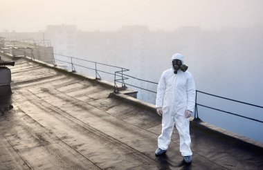 Beyaz tulumlu ve gaz maskeli bir adam sisli havada yükselen güneşe arkası dönük bir şehir binasının çatısında dikiliyor. Çevresel sorun kavramı.