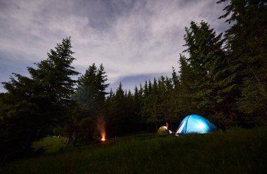 Akşamları ormanda kamp yapmak. Çadırlar ve yanan şenlik ateşi yıldızlı gökyüzünün altındaki yeşil köknar ağaçlarının arasında dumanla birlikte. Lambaların parladığı çadırların arasındaki kişi. Sağlıklı yaşam tarzı kavramı