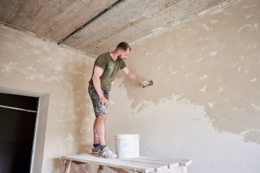 Erkek müteahhit ahşap bir stantta duruyor ve duvarında alçı olan bir spatulayla küçük bir odada çalışıyor. Duvarları içeri koyuyoruz. Tişörtlü ve kotlu sakallı işçi boyalı.
