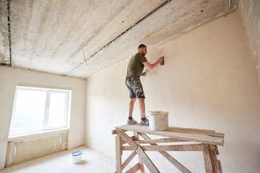 İnşaatçı adam ahşap bir standın üzerinde duruyor ve duvarında alçı olan bir spatulayla pencereye karşı çalışıyor. Duvarları içeri koyuyoruz. Tişört ve kot giyen sakallı bir adam boyayla boyanmış.