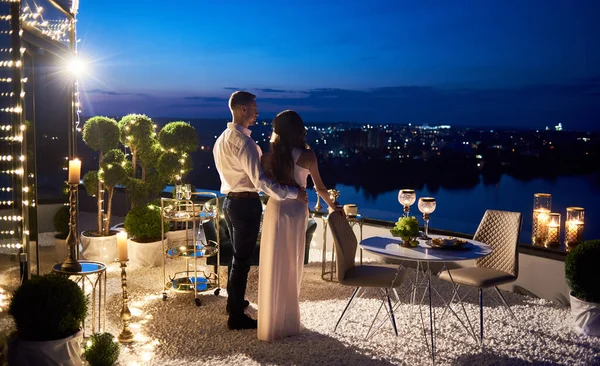 Back view of beautiful couple in love having romantic date at rooftop restaurant. Stylish man hugging charming girlfriend and looking at magnificent night cityscape. Concept of love and romance.