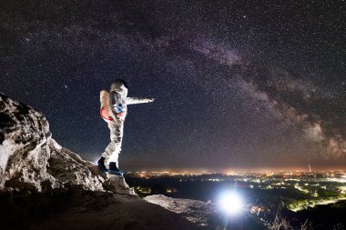 Uzay yolcusu yıldızlarla dolu görkemli gökyüzünün altında kayalık tepede dururken gece şehrini işaret ediyor. Görev uzmanı astronot beyaz uzay giysisi giyiyor. İnsan uzay keşfi kavramı.