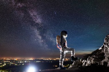 Uzay yolcusu yıldızlarla dolu karanlık gökyüzünün altında kayalık tepede duruyor. Görev uzmanı astronot kasklı beyaz bir uzay giysisi giyiyor. Kozmonotlar, Samanyolu ve uzay yolculuğu kavramı.