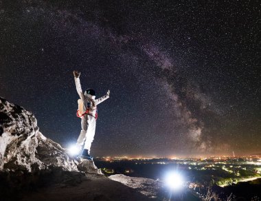 Astronot, arka planda Samanyolu ile muhteşem yıldızlı gökyüzü ile kayalık dağda dururken kollarını kaldırıyor. Uzay adamı beyaz uzay giysisi ve miğfer giyiyor. Uzay yolculuğu kavramı