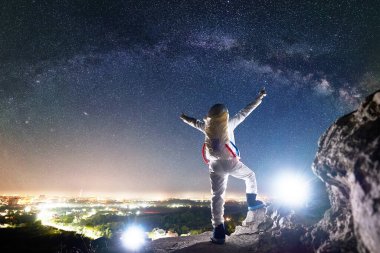 Arkaya bakan uzay yolcusu, yıldızlı güzel gökyüzünün altında kayalık tepede dururken kollarını yana doğru açıyor. Uzay giysisi konusunda uzman astronot Samanyolu, Gece Şehri manzarasının tadını çıkarıyor.