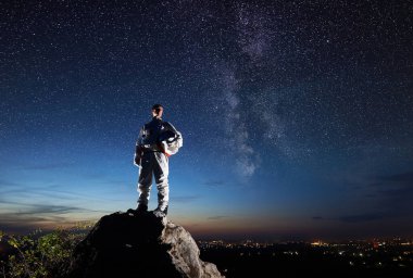 Uzay yolcusu Rocky Hill 'in tepesinde duruyor ve yıldızlı güzel gökyüzüne bakıyor. Beyaz uzay giysili astronot miğfer tutuyor. Uzay yolculuğu ve kozmonotik kavramı.