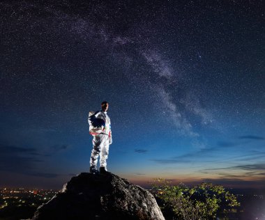 Kayalık tepenin üstünde duran ve yıldızlarla dolu güzel gökyüzüne bakan uzay yolcusunun yan görüntüsü. Beyaz uzay giysili astronot miğfer tutuyor. Uzay yolculuğu ve Samanyolu kavramı.