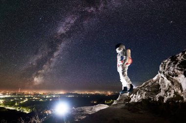 Gece gökyüzünün altındaki kayalık dağda yıldızlarla birlikte duran cesur uzay yolcusunun yan görüntüsü. Uzay giysisi konusunda uzman astronot, gece şehri manzarasının keyfini çıkarıyor. Samanyolu ve uzay yolculuğu kavramı.