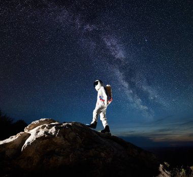 Uzay yolcusu yıldızlarla ve Samanyolu 'yla nefes kesici gece gökyüzü altında kayalık dağa tırmanıyor. Uzay giysisindeki astronot dev bir kayanın tepesine ulaşıyor. Uzay keşfi ve kozmonotik kavramı.