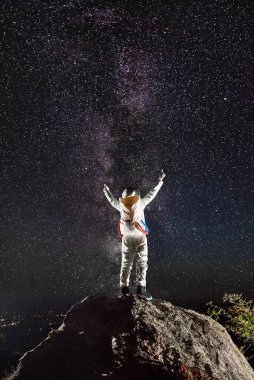 Cesur kozmonotun dağın tepesinde dikilirken kollarını kaldırışının arkası. Uzay kıyafetli astronot yıldızlı güzel gece gökyüzünü seyrediyor. Uzay yolculuğu kavramı, kozmonotik