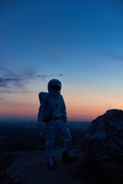 Gün batımında kayalık dağda duran astronot siluetinin dikey görüntüsü. Kozmonot görkemli gökyüzüne bakarken beyaz uzay elbisesi giyiyor. Uzayda insan kavramı.
