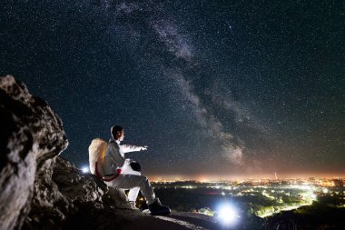 Yıldızlı gökyüzünün altında, Samanyolu kayalık dağda otururken, uzay adamının vadideki gece şehrini işaret edişinin yan görüntüsü. Uzay elbisesi giymiş uzay yolcusu miğfer tutuyor. Uzay yolculuğu kavramı