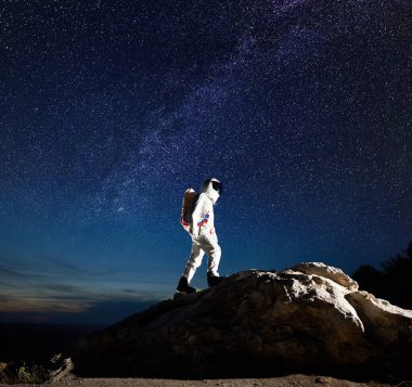 Uzay yolcusu, yıldızlarla nefes kesen gece gökyüzü altında kayalık dağa tırmanıyor. Uzay giysisindeki astronot dev bir kayanın tepesine ulaşıyor. Samanyolu ve uzay keşfi kavramı.