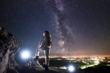Uzay yolcusunun kayalık dağda dikilip yıldızlı gökyüzünün, Samanyolu 'nun ve gece şehrinin tadını çıkarışının arkası. Görev uzmanı astronot beyaz uzay giysisi giyiyor. Uzay yolculuğu kavramı