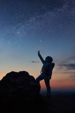 Uzay yolcusu kayalık dağda duruyor ve yıldızları işaret ediyor. Silüet astronot, yıldızlı gökyüzüne bakarken beyaz uzay elbisesi giyiyor. Galaksi kavramı, uzayda insan ve samanyolu..
