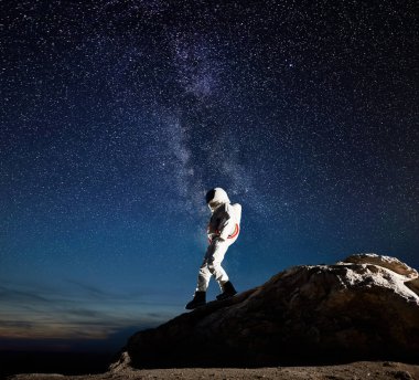 Yıldızlarla dolu muhteşem gece gökyüzü altında kayalık tepeden aşağıya doğru yürüyen bir astronot. Uzay giysisi içindeki kozmonot yeni gezegenin yüzeyini keşfediyor. Galaksi, Samanyolu ve uzay yolculuğu kavramı.