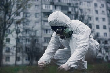 Çömelmiş ekolojist, laboratuvar aletleri ve sıvılarla yeni numunelerle çalışıyor. Gezegenimizi kurtarma kavramı. Beyaz üniformalı, eldivenli ve gaz maskeli bir işçi.