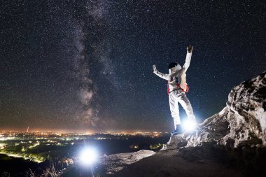 Uzay yolcusu tam uzunlukta kollarını kaldırırken kayalık dağda fantastik yıldızlı gökyüzü, arka planda Samanyolu. Uzay adamı beyaz uzay giysisi ve miğfer giyiyor. Uzay yolculuğu kavramı