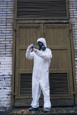 Eski kapının önünde dikilip mavi sıvı laboratuvar deneyiyle cam şişedeki tepkiyi gözlemleyen ekolojistin tam boy görüntüsü. Beyaz üniformalı, eldivenli ve gaz maskeli bir bilim adamı.
