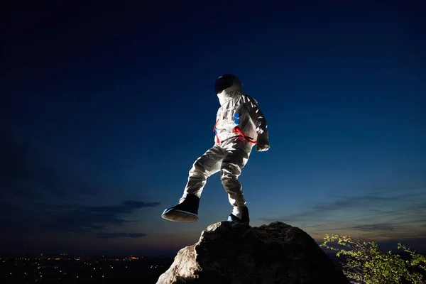 Full length of astronaut standing on one leg on top of mountain with beautiful night sky on background. Cosmonaut wearing white space suit with helmet. Concept of space travel and cosmonautics.