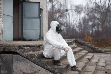 Koruyucu üniforma ve gaz maskesi takan bilim adamı, eski bir binanın merdivenlerinde oturuyor, başka tarafa bakıyor, arka plandaki kapıları açıyor.