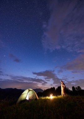 Erkek turist, yıldızlarla dolu güzel bir gökyüzünün altında, aydınlık bir çadır ve kamp ateşiyle akşamları dağlarda vakit geçirir. Yazın dağlarda gece kampı.