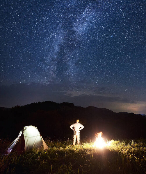 Rear view of man tourist near illuminated tent and campfire enjoying starry sky, Milky way against background of mountains. Beautiful landscape of the night sky with bright stars and clouds appearing