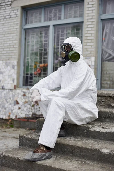 古い建物のドアの上に座ってガスマスクの男性環境保護主義者 白い保護制服 靴カバー 手袋を着用する研究科学者 生態系と環境汚染の概念 — ストック写真