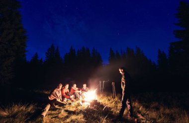 Yürüyüşçüler, ormanda kamp alanında ateş yakarak dinleniyorlar. Turistler, güçlü köknar ağaçlarının ve yıldızların göründüğü gece gökyüzünün zemininde kütüğün üzerinde oturuyorlar.