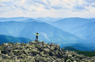 Arkaya bakan kadın kollarını açıyor, uçurumun tepesinde güçlü bir dağ vadisiyle dikiliyor. Parlak ceketli, uzun saçlı bir bayan turist bulutlu bir günde manzaranın tadını çıkarıyor..