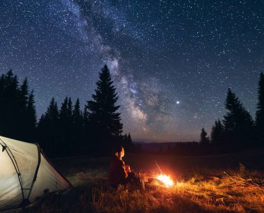 Güneşli gökyüzü ve parlak Samanyolu 'nun altında güzel bir kamp gecesinin tadını çıkaran, çadırın yanında yanan parlak kamp ateşinin yanında tek başına oturan kadının arkası. Dağlarda sıcak bir yaz akşamı..