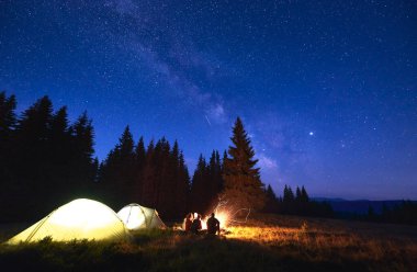 Şenlik ateşiyle gece kampı. İnsanlar çadır kenti yakınlarında kamp ateşinde güneşleniyor, çam ormanlarındaki dağ vadisinin tadını çıkarıyorlar. Koyu mavi gece gökyüzü parlak yıldızlar ve Samanyolu ile kaplı.