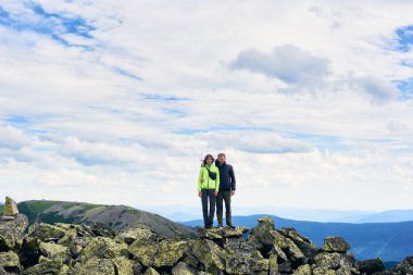 Sevgi dolu bir çiftin kucaklaşması, poz vermesi ve kameraya bakması, arka planda güzel bir dağ manzarası. Dağ yürüyüşü, turistler birlikte zirveye ulaşıyor. Vahşi doğa, spor turizmi konsepti.