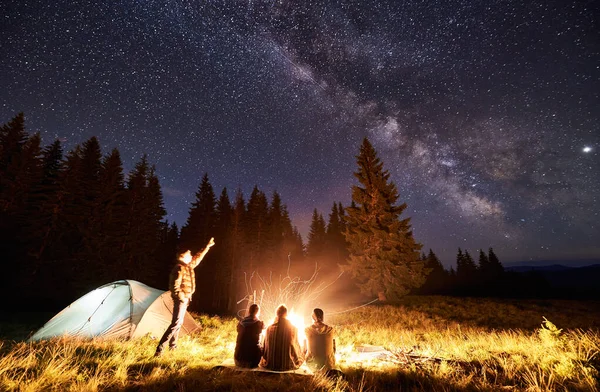 Male showing his friends Milky Way over camping. Guys are sitting by the campfire on the background of the valley of mountains and pine forest, enjoying the evening starry sky. Outdoor recreation