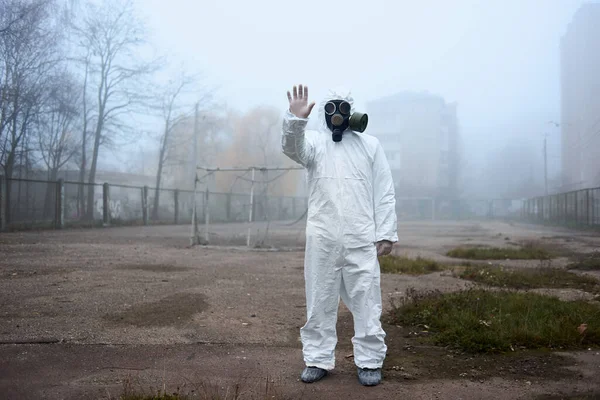 Man in gas mask and protective costume showing stop gesture with hand up.