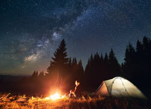 Night camping in the mountains. Young woman hiker sitting by campfire and illuminated tourist tent. On background spruce forest under beautiful night starry sky with Milky way. Concept of tourism