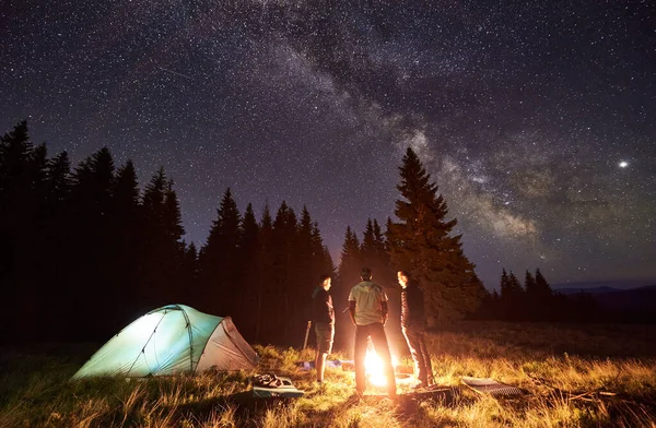 Night summer camping in forest. Bright campfire burning, three male tourists standing around fire near tent under beautiful dark starry sky and Milky way. Concept of tourism, camping in the mountains.