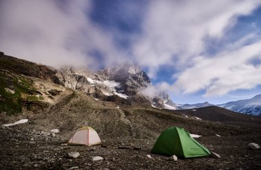 Gece dağlarında kamp yaparken panoramik manzara. Kayalık tepe ve dağ tepelerindeki turist çadırlarının manzarası yıldızlı, sisli gökyüzünün altında. Seyahat, yürüyüş ve kamp kavramı.