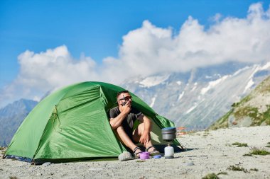 Kamp çadırında oturan ve telsiz kullanan bir turist. Elinde radyosu olan, dağları ve mavi gökyüzü olan turist çadırında dinlenen bir erkek gezgin. Seyahat, yürüyüş, kamp yapma kavramı
