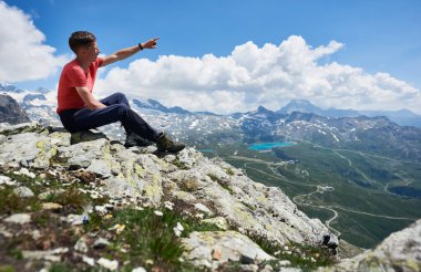 Tişörtlü turist güzel dağ manzarasına bakıyor ve parmağıyla işaret ediyor. Zirveye ulaşan, taşların üzerinde oturan adam. Güneşli yaz gününde harika manzaralı vahşi doğa. Alplerde spor turizmi.