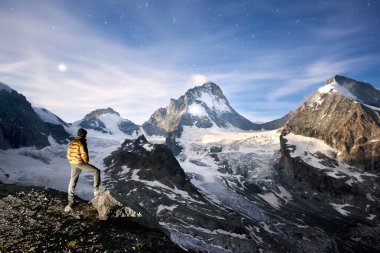 Yıldızlarla dolu inanılmaz mavi gökyüzünün yatay görüntüsü ve İsviçre 'deki Pennine Alpleri' nin soğuk ve karlı zirveleri üzerinde Ay. Bir turist durup güzelliği izliyor.