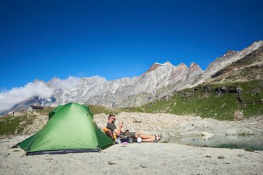 Arka planında görkemli kayalık tepeler olan turist çadırının yanında oturan erkek turistin yan görüntüsü. Akıllı telefon kullanan ve mavi gökyüzünün altındaki dağlarda dinlenen bir adam. Seyahat, yürüyüş ve kamp kavramı