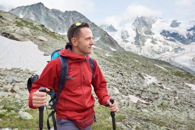Kırmızı ceketli ve sırt çantalı bir adam portresi, İsviçre Alpleri 'ndeki dağlarda yürüyüş direkleri ile yürüyüş yapmak, etrafındaki muhteşem manzara.