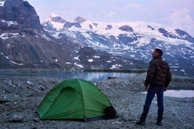 Manzaranın tadını çıkaran bir adamın arka görüntüsü, boş kayalık dağlarda çadırın yanında duruyor, temiz suyu olan küçük göllerin yanında. Popüler turist eğlencesi. Doğa güzelliği kavramı.