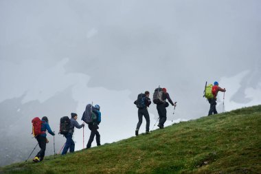 Otlu tepeye tırmanırken sırt çantalı yürüyüşçülerin arka planı. Gençler arka planda sisli uçurumla dağlarda yokuş yukarı yürüyorlar. Yürüyüş, seyahat ve turizm kavramı.