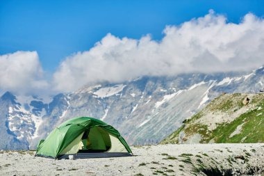 Dağ vadisinde yeşil turist çadırı, muhteşem kayalık tepeler ve arka planda mavi gökyüzü. Dağ manzarası, dağ sığınağı, tepenin üstündeki beyaz bulutlar. Seyahat, kamp ve yürüyüş kavramı.